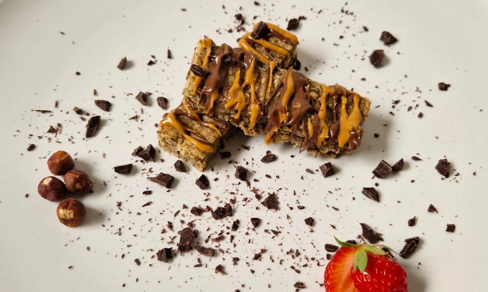 zwei Proteinriegel mit Erdnussbutter und Schoko-Topping, daneben Schokosplitter, Nüsse und eine Erdbeere als Deko