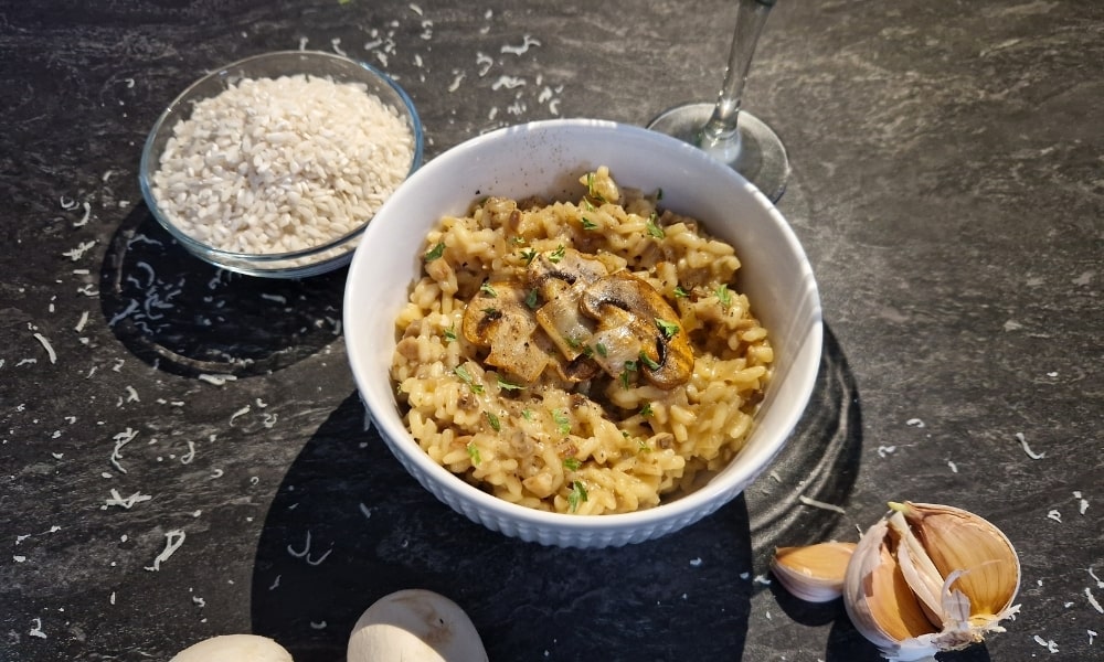 Schüssel mit veganem Pilzrisotto, daneben roher Risottoreis, Knoblauch und ein Glas Wein