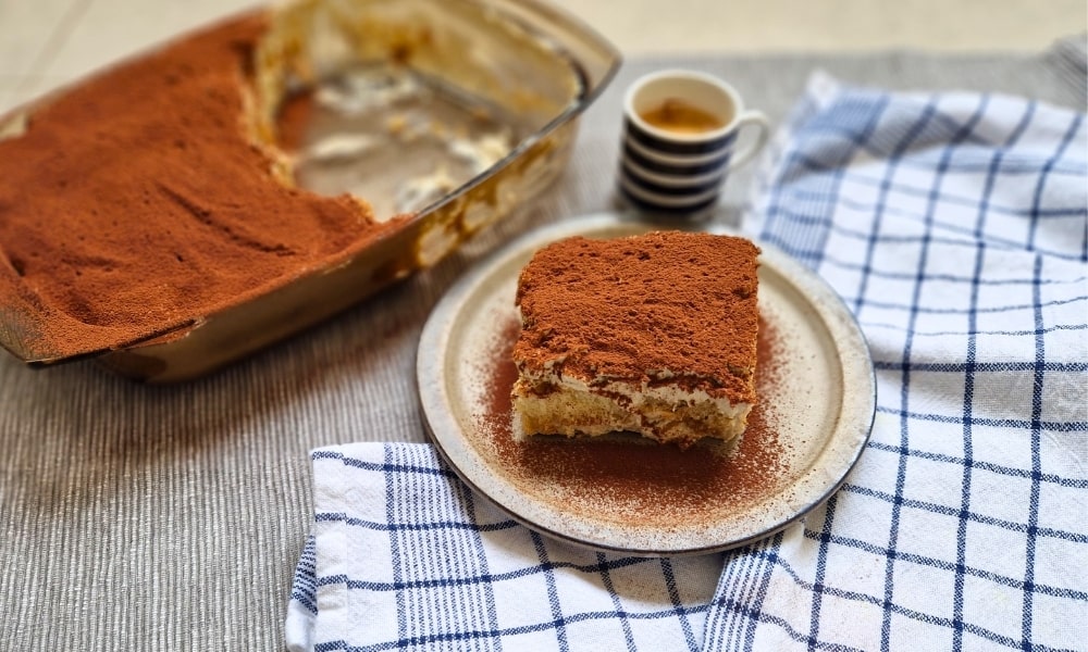 cremiges italienisches Protein Tiramisu auf einem Teller mit einem Espresso, im Hintergrund eine Glasschale mit mehr Tiramisu