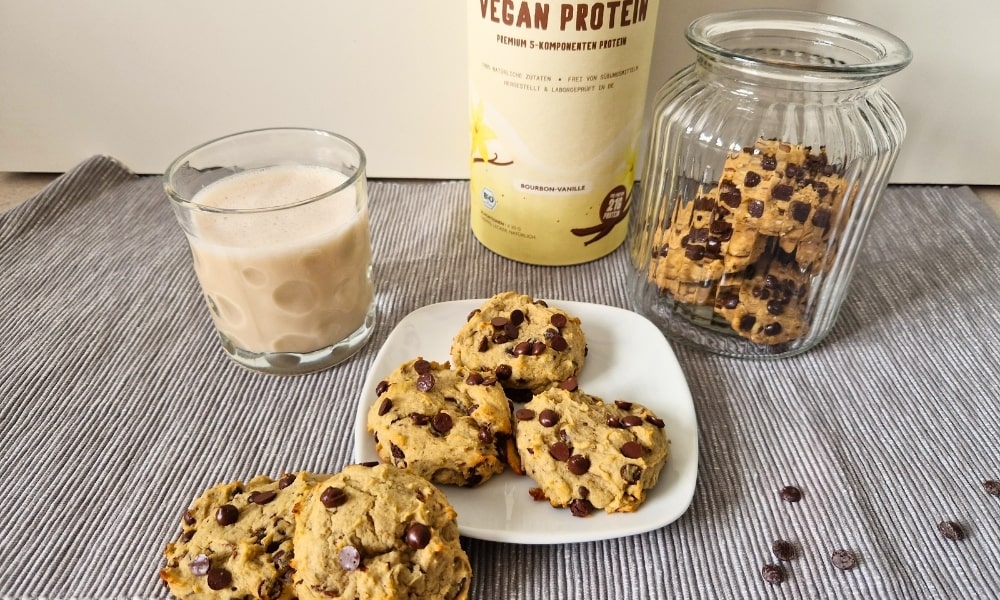 Protein Cookies auf einem Teller mit einem Glas Milch, Vanille Proteinpulver von Vetain und einem Glas mit weiteren Cookies im Hintergrund