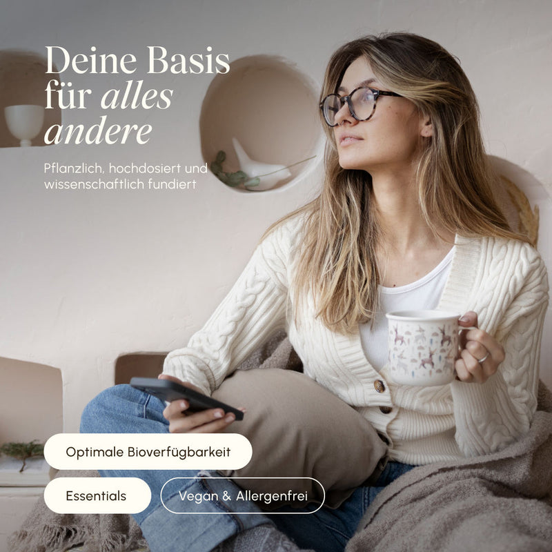 Woman with glasses sitting relaxed on the sofa with a cup of tea and smartphone. Text: "Your foundation for everything else – plant-based, high-dose, and scientifically proven." Badges: Optimal bioavailability, Essentials, Vegan & Allergen-free