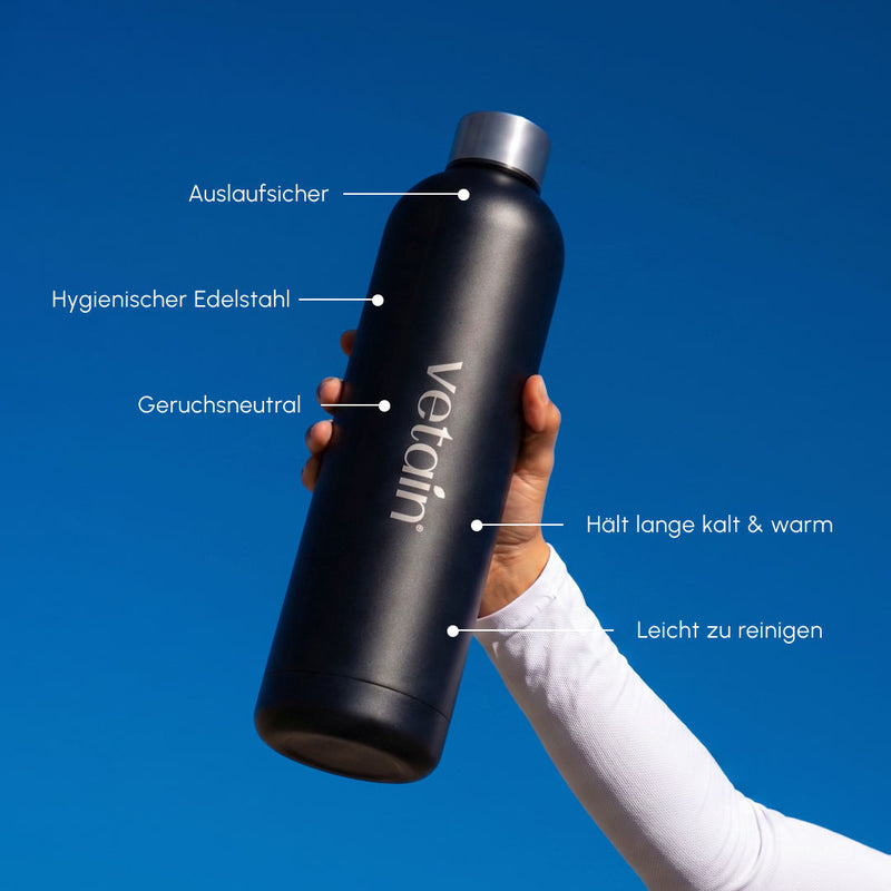 Hand hält eine schwarze vetain Edelstahl-Trinkflasche vor blauem Himmel; Beschriftungen nennen auslaufsicher, hygienischer Edelstahl, geruchsneutral, hält lange kalt und warm sowie leicht zu reinigen.