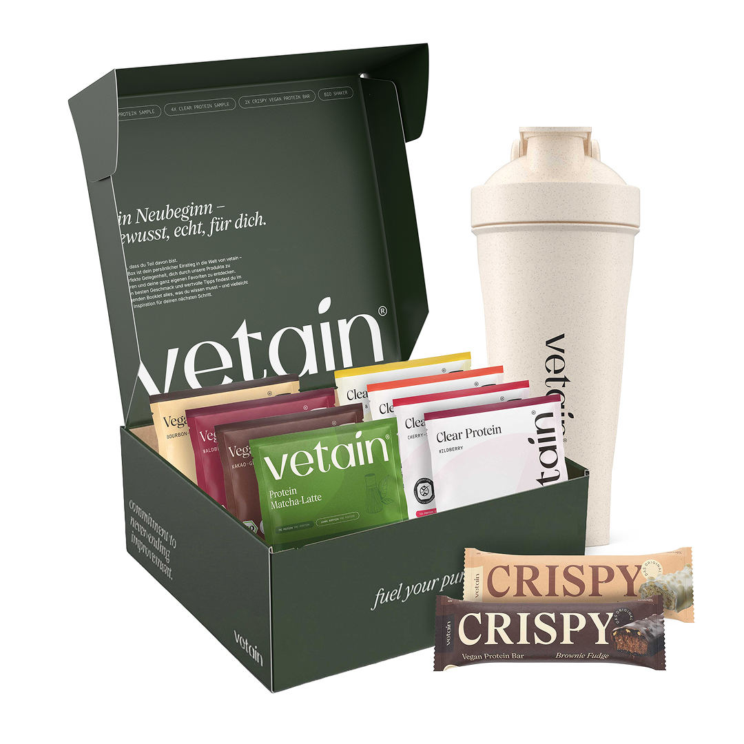 Geöffnete grüne vetain Probierbox mit Innendeckel-Text „Ein Neubeginn – bewusst, echt, für dich.", gefüllt mit Vegan- und Clear-Protein-Proben, daneben ein beiger vetain Shaker und zwei CRISPY Proteinriegel in den Sorten Zimtschnecke und Brownie Fudge.