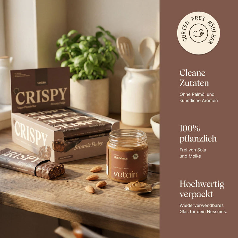 Vetain Crispy Vegan Protein Bar in der Sorte Brownie Fudge (Box und angeschnittener Riegel) neben einem Glas Bio-Mandelmus auf einer Küchentheke. Badge „Flavours selectable“. Hinweise: clean ingredients (ohne Palmöl und künstliche Aromen), 100 % plant-based (frei von Soja und Molke), hochwertige, wiederverwendbare Glasverpackung.